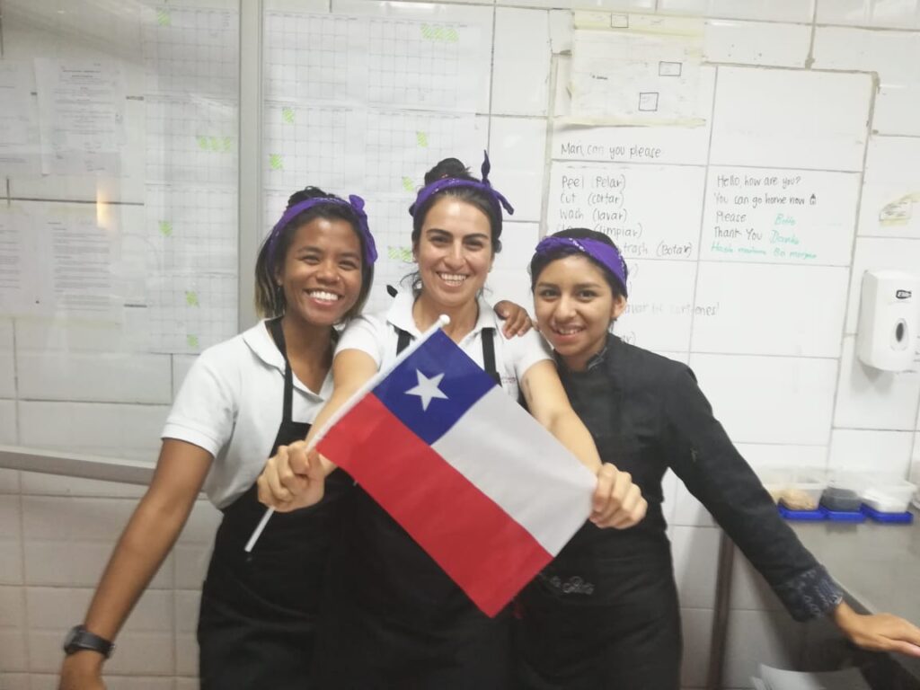 Marielle standing in a restaurant kitchen where she worked with two of her colleagues who are holding. Chilean flag.